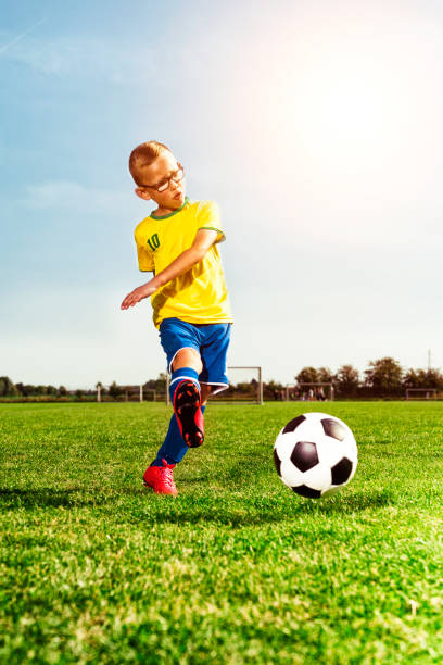 One boy with glasses wears soccer clothes and kicks a ball. The boy runs on a soccer field. The sun shines. It is summer.