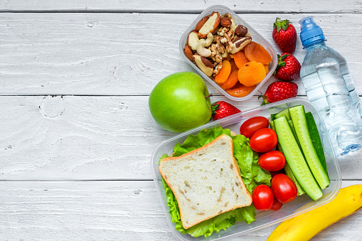 school lunch boxes with sandwich and fresh vegetables, bottle of water, nuts and fruits on white wooden background. healthy eating concept. top view with copy space