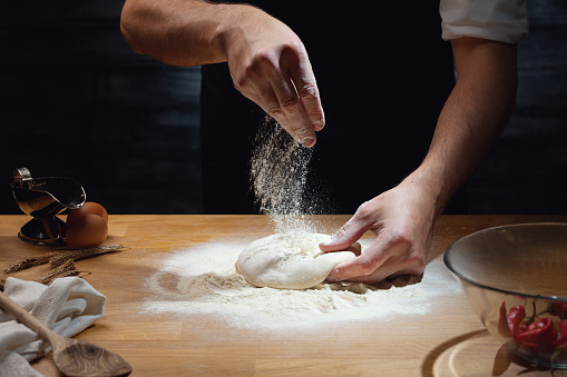 Cook hands kneading dough, sprinkling piece of dough with white wheat flour. Low key shot, close up on hands, some ingredients around on table.