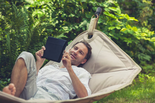 man in a hammock using digital tablet man resting out in the garden in a hammock and using his digital tablet. hammock men lying down digital tablet stock pictures, royalty-free photos & images