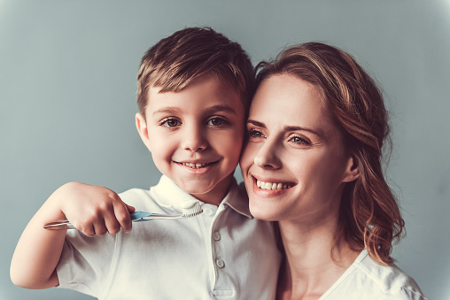 Beautiful woman and her cute little son are hugging and smiling, on gray background. Boy is holding a toothbrush and looking at camera