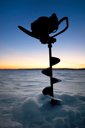 Ice auger standing up in a partially drilled hole awaiting the next ice fishing hole to be drilled, during the last light of the day