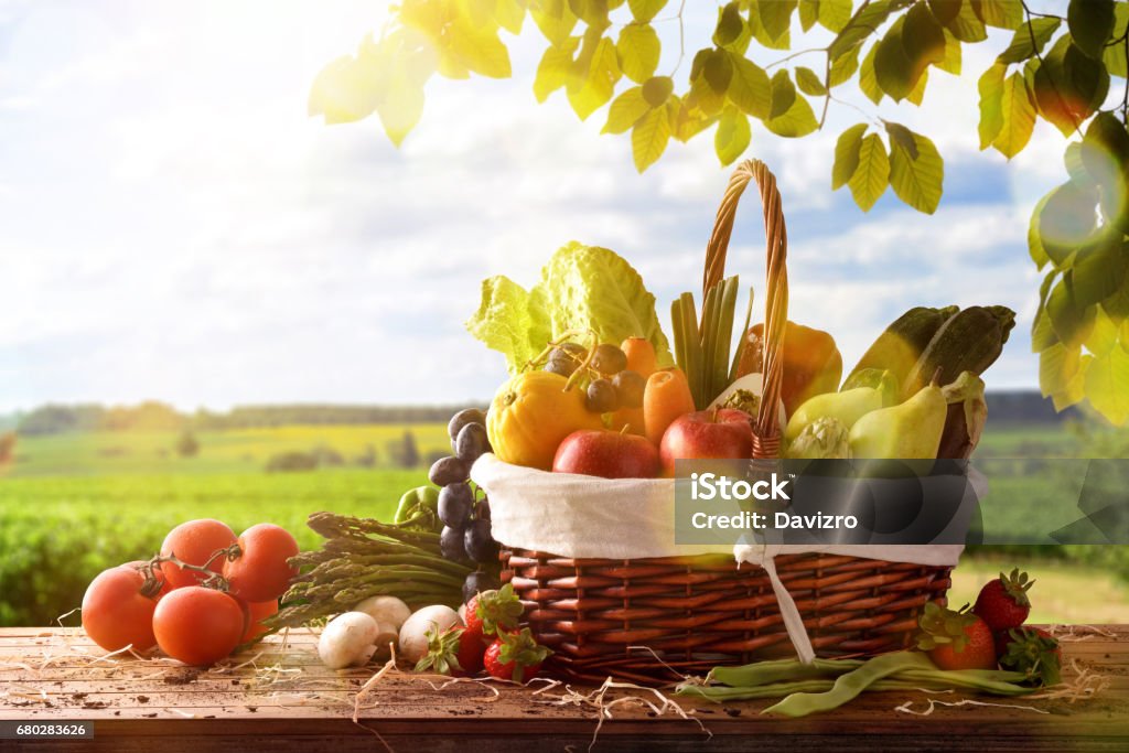 Fruits and vegetables on table and crop landscape background Assortment of fruits and vegetables in a wicker basket on a wooden table on crop landscape background. Horizontal composition. Front view Fruit Stock Photo