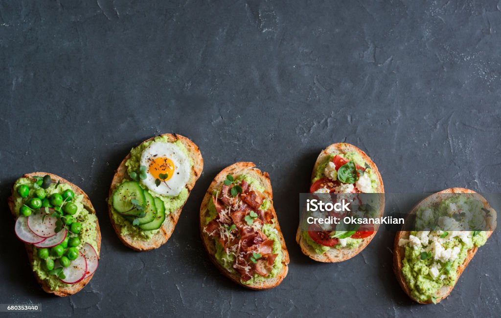Breakfast avocado sandwich - with crispy bacon, quail egg, tomatoes, goat cheese, green peas, radish, cucumber. Healthy snack. On a dark background, top view, copy space Avocado Stock Photo