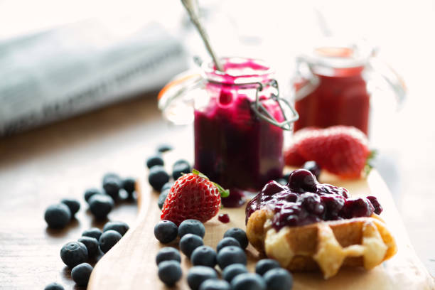 Messy Breakfast, Blueberry Jam, Mini Belgian Waffles, Fresh Strawberries, Newspaper Breakfast of Belgium Liege waffles with strawberry and blueberry compote, sauce or jam spooned from opened mason jars.  Fresh blueberries and strawberries on the side.  All set on a wood cutting board.  Drips, spills and a little messy.  Closeup view.

 compote photos stock pictures, royalty-free photos & images