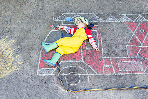 Creative leisure for kids: adorable toddler child  having fun with fire truck picture drawing with chalk, outdoors. Dreaming of future profession.