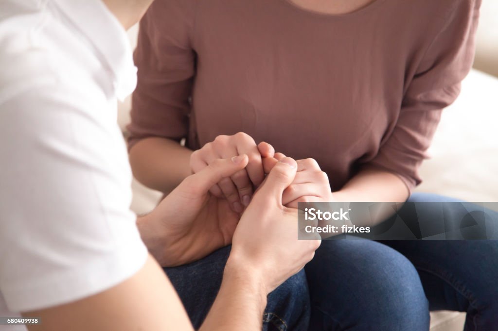 Close up of man holding hands of woman, proposal, apologize Close up view of man holding hands of a woman sitting on sofa indoors, love confession or marriage proposal, expressing support and comforting, asking for forgiveness and apologizing Embracing Stock Photo
