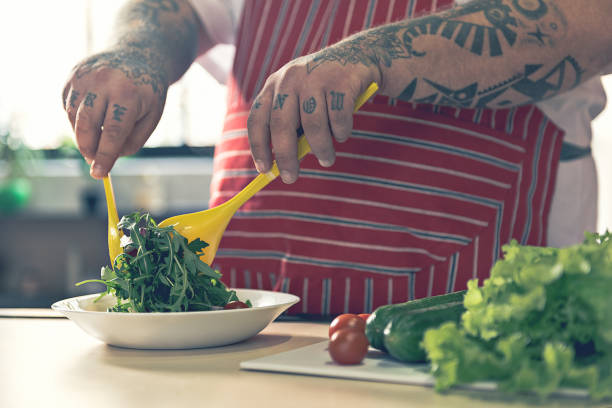 Close up of male hands with tattoo preparing healthy food in kitchen. Thick guy is mixing chopped vegetables in plate