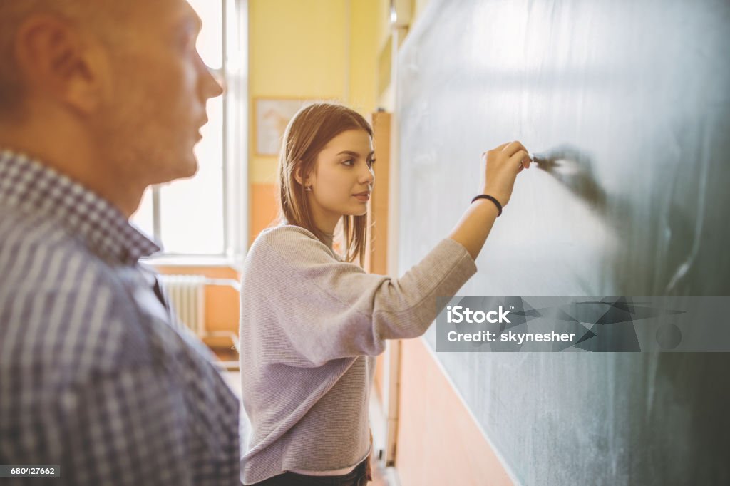 Female high school student writing on blackboard in the classroom. Female teenage student writing on blackboard during the class while teacher is standing next to her. Chalkboard - Visual Aid Stock Photo