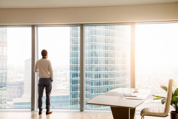 jeune homme d’affaires prospère debout en fonction regardant la fenêtre pleine longueur - rear view businessman thinking men photos et images de collection