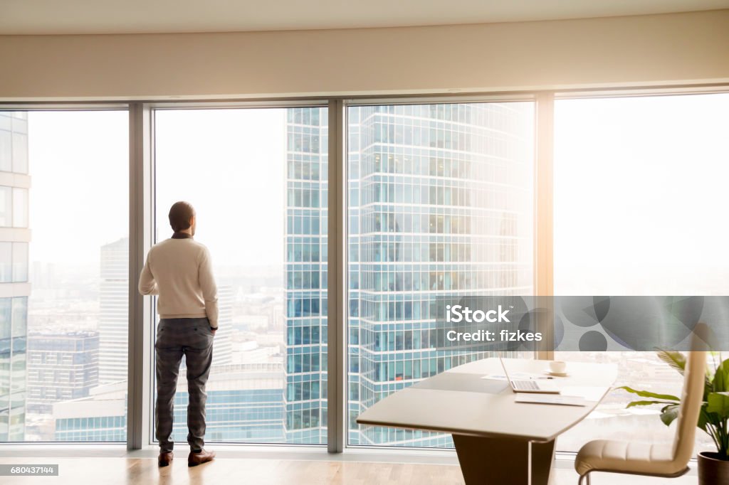 Young successful businessman standing in office looking at full-length window Rear view at young successful businessman standing in office looking through full-length window at cityscape with skyscrapers, dreaming or resting, waiting for meeting or considering business offer Luxury Stock Photo