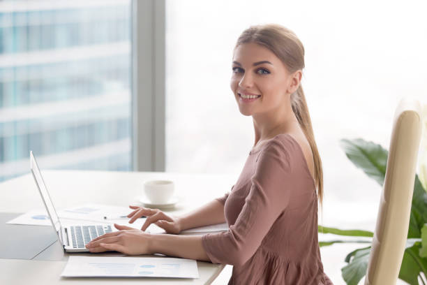 jeune femme souriante, assis au bureau en regardant la caméra - niveau junior photos et images de collection