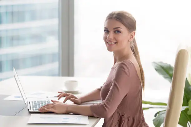 Portrait of smiling young attractive manager, happy businesswoman sitting at workplace using laptop and looking at camera. Starting career in big company, accounting services and business analysis