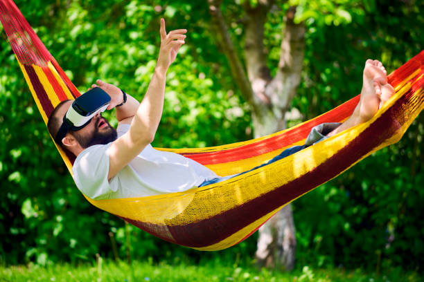 istock Young man wearing virtual reality goggles relaxing in garden hammock. 680450014