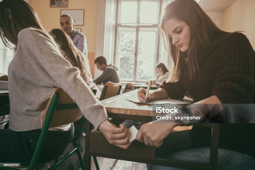 Two high school students passing note during the exam in the classroom. Teenage girls cheating during exam and passing paper note in the classroom. Dishonesty Stock Photo