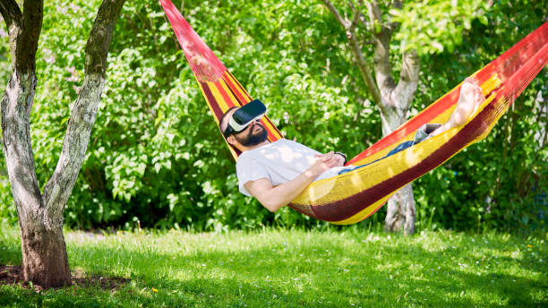 istock Young man wearing virtual reality goggles relaxing in garden hammock. 680454434