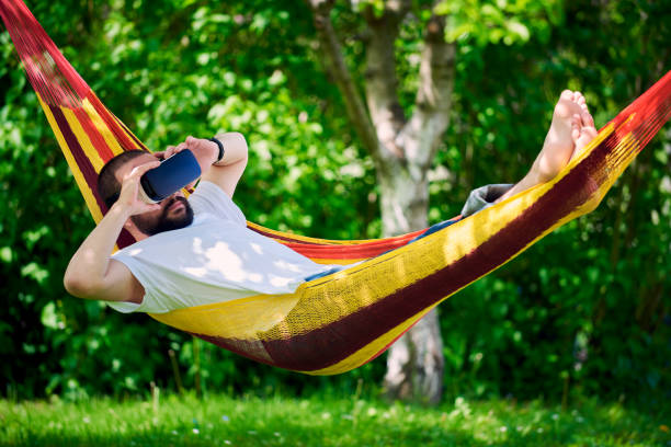 istock Young man wearing virtual reality goggles relaxing in garden hammock. 680460712