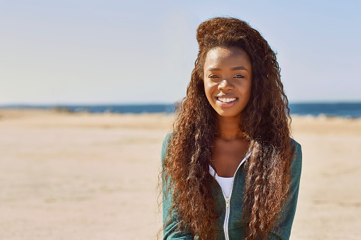 Portrait of young woman smiling at beach. Fit female is with long curly hair. She is in sportswear on sunny day.