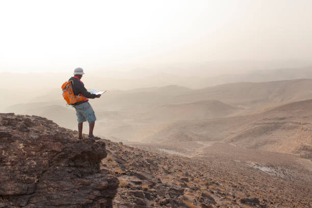 Outdoor active man standing on desert rocky mountain peak, reading trekking map. Backpacker tourist wearing long shorts, outdoor sandals, bucket hat and  orange backpack.