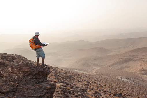 Outdoor active man standing on desert rocky mountain peak, reading trekking map. Backpacker tourist wearing long shorts, outdoor sandals, bucket hat and  orange backpack.