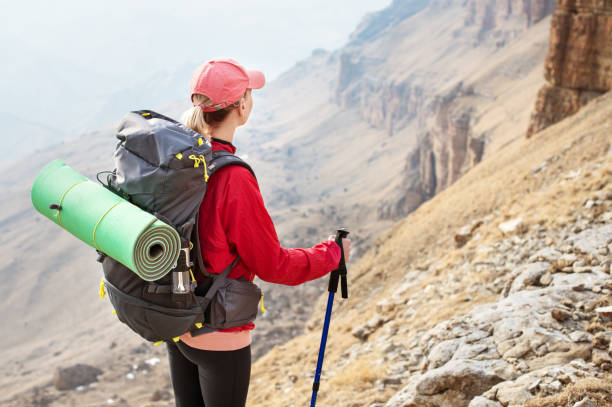 istock A slender girl in a cap with sticks for Nordic walking with a backpack and a folded rug for relaxation stands in the mountains against the backdrop of rocks and distant lands 680494068