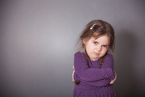 A little cute Girl is sulking. She is wearing a purple shirt in front of a gray background.