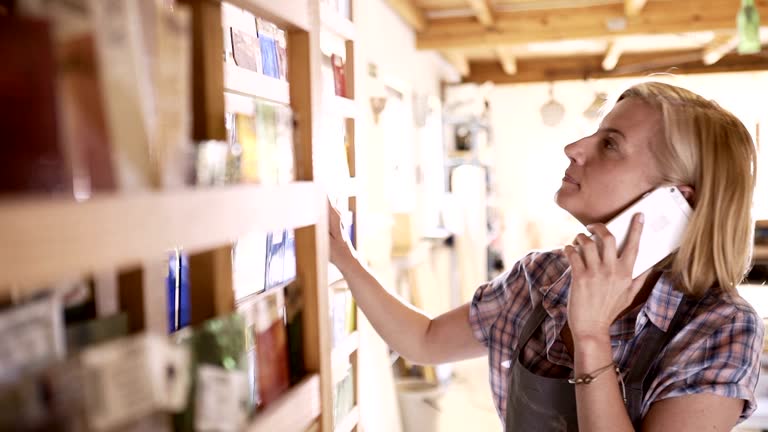 Female artist talking on cell phone and viewing stained glass samples on shelves in studio