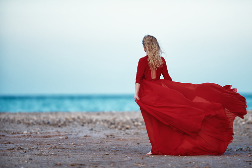 rear view of blond hair woman wearing red long dress, posing on the beach and enjoying the silence moment.freedom concept.