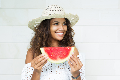 [Note for inspector: Photo files for becoming exclusive contributor] Closeup portrait of smiling young beautiful woman holding slice of watermelon with tile wall in background. Front view.