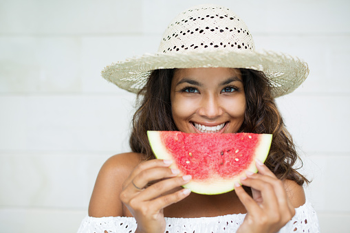 [Note for inspector: Photo files for becoming exclusive contributor] Closeup portrait of smiling young beautiful woman looking at camera, holding slice of watermelon and eating it. Front view.