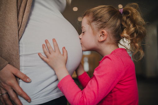 A little girl is kissing her mothers pregnant belly and smiling happily in anticipation.