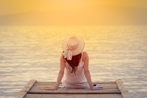 Woman in hat sitting at the end of dock and enjoying the sea view. Summer vacation concept.