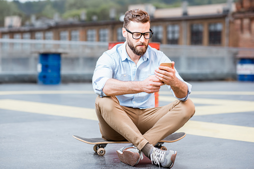 Hipster businessman sitting with smart phone on the rooftop playground of the industrial building. Lifestyle business concept