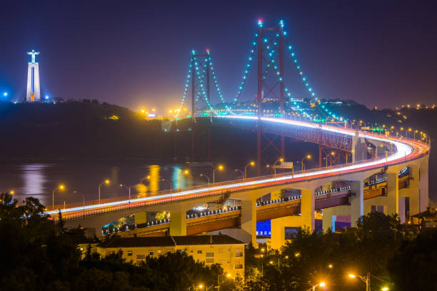 The iconic span of the 25 de Abril Bridge illuminated by the lights of the suspension cables and the zooming traffic crossing from Almada to Lisbon, Portugal’s vibrant capital city.