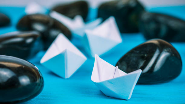 istock Teamwork of white paper ships between abstract rock stones on blue background 680775558