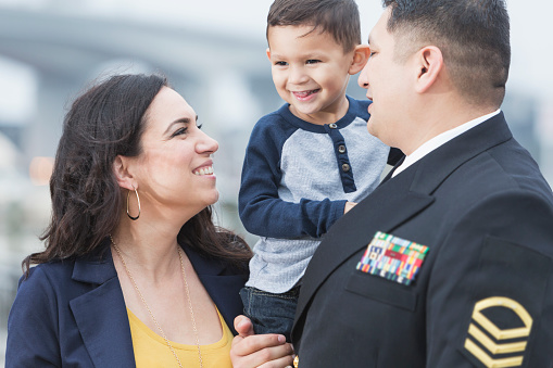 An Hispanic man wearing a military uniform, standing with family. He is holding his smiling 2 year old son, and standing beside his happy wife.