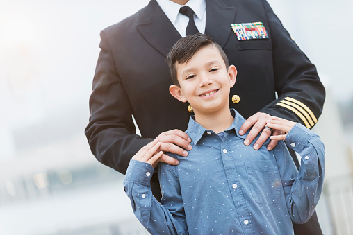 Cropped view of an Hispanic navy officer standing behind his  8 year old son. The boy is smiling and touching his dad's hands, which are on his shoulders.