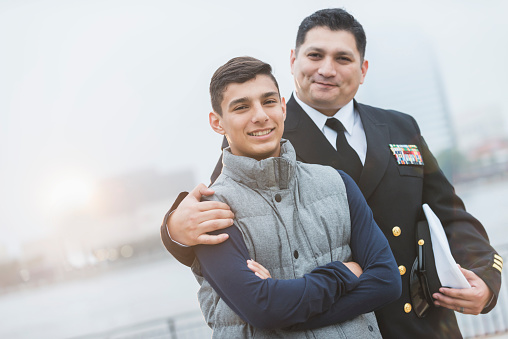 An Hispanic man wearing a military uniform, standing with his teenage son. The 13 year old boy is looking smiling at the camera.