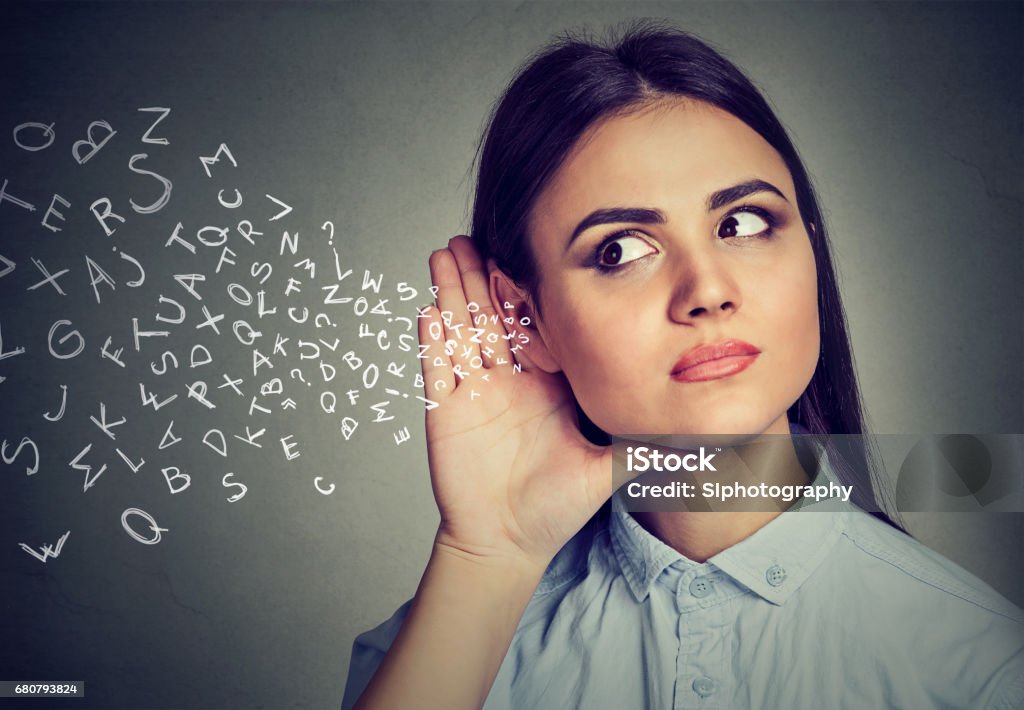 Woman holds hand near ear and listens carefully alphabet letters flying in Woman holds her hand near ear and listens carefully alphabet letters flying in isolated on gray wall background Listening Stock Photo