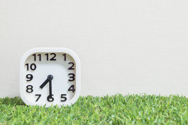 istock Closeup white clock for decorate show a half past seven or 7:30 a.m. on green artificial grass floor and cream wallpaper textured background with copy space 680819228