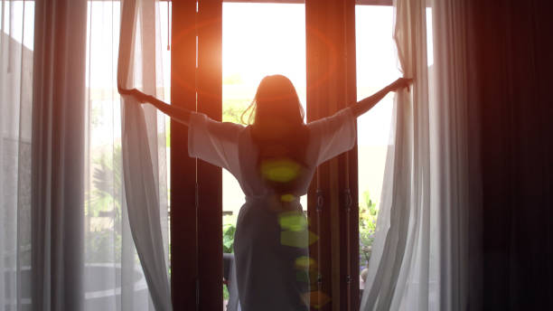 mujer joven en bata de baño abra cortinas y estirar de pie cerca de la ventana en su casa. - sun watch fotografías e imágenes de stock