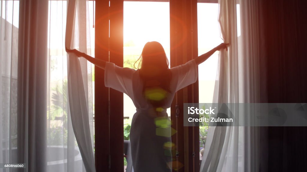 Mulher jovem em roupão abrir cortinas e esticar em pé perto da janela em casa. - Foto de stock de Hotel royalty-free