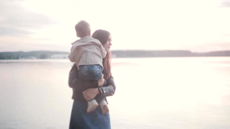 Mother holding son on her hand on the shore of the beach. Smiling woman pick up her little boy, kiss and hug