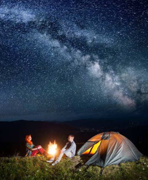 istock Night camping. Happy couple backpackers sitting by bonfire and tent under incredibly beautiful starry sky. Village in the valley on the background. Long exposure 680865254