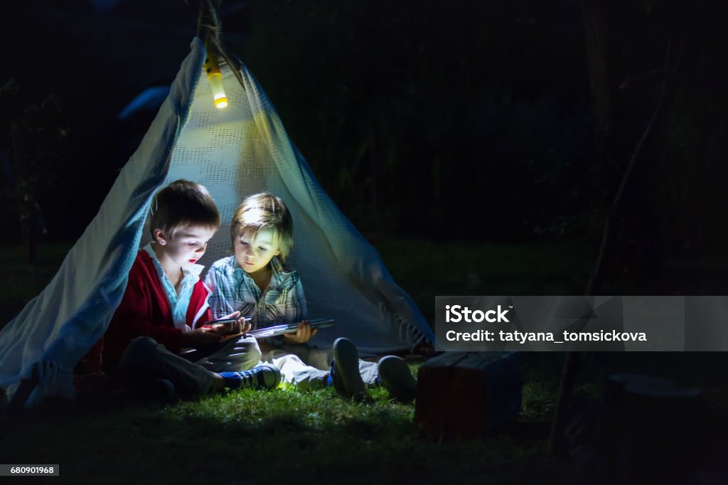 Cute little brothers, playing on tablet and telephone at night in campside, in the tent Camping Stock Photo
