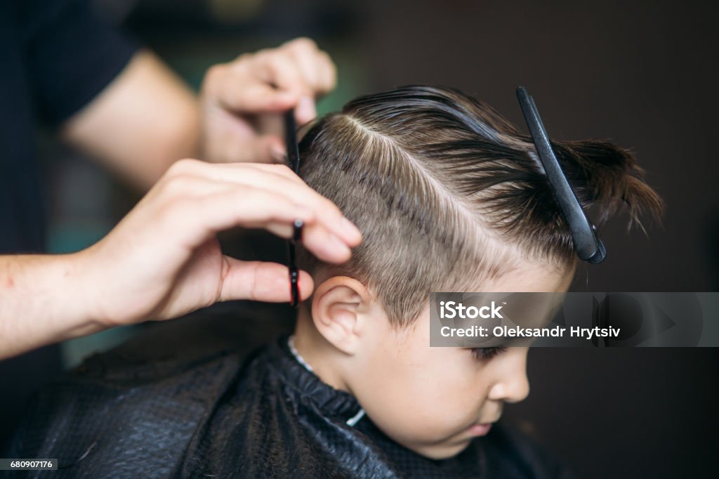 Little Boy Getting Haircut By Barber While Sitting In Chair At Barbershop. Little boy on a haircut in the barber sits on a chair. Child Stock Photo