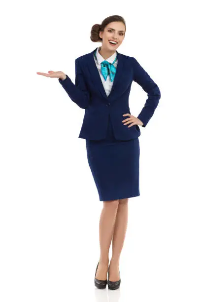 Smiling young businesswoman in blue formalwear and high heels is standing with hand raised and looking at camera. Front view. Full length studio shot isolated on white.