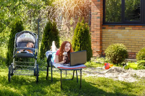 Photo of Young woman with her newborn baby lying on the soft armchair with laptop. Summer day. Mother with child outdoors. Motherhood.