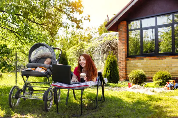 Photo of Young woman with her newborn baby lying on the soft armchair with laptop. Summer day. Mother with child outdoors. Motherhood.