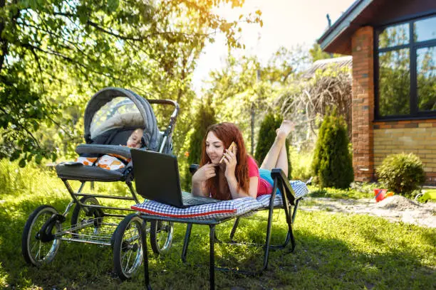 Photo of Young woman with her newborn baby lying on the soft armchair with laptop and talking on phone. Summer day. Mother with child outdoors. Motherhood.